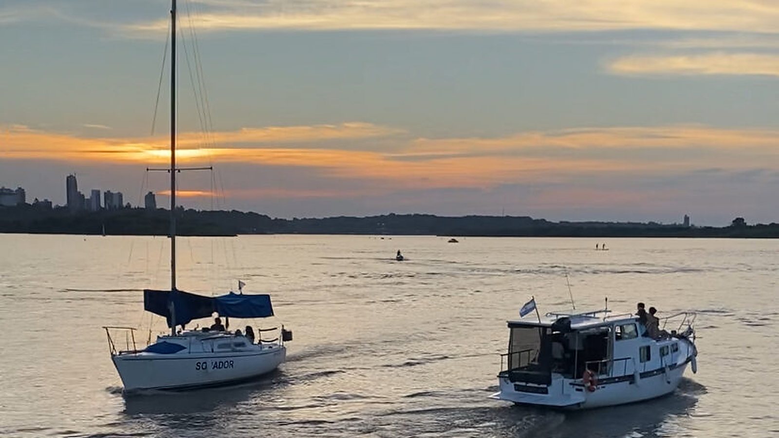Velero y lancha navegando al atardecer frente al skyline de Paraná